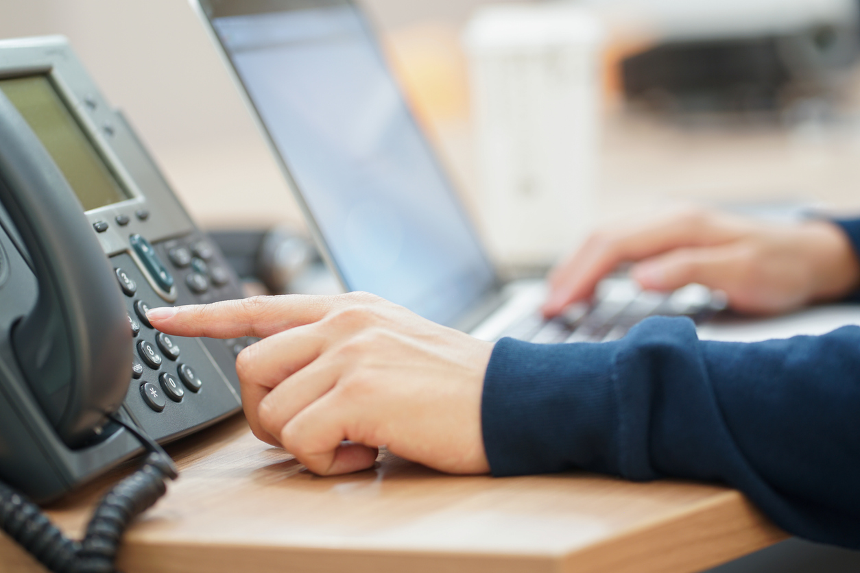 A person's hand pushing a button on a VoIP phone at a desk with a laptop nearby.