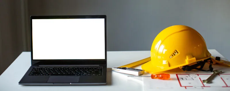 A laptop, yellow hard hat, and blueprints on a construction office desk